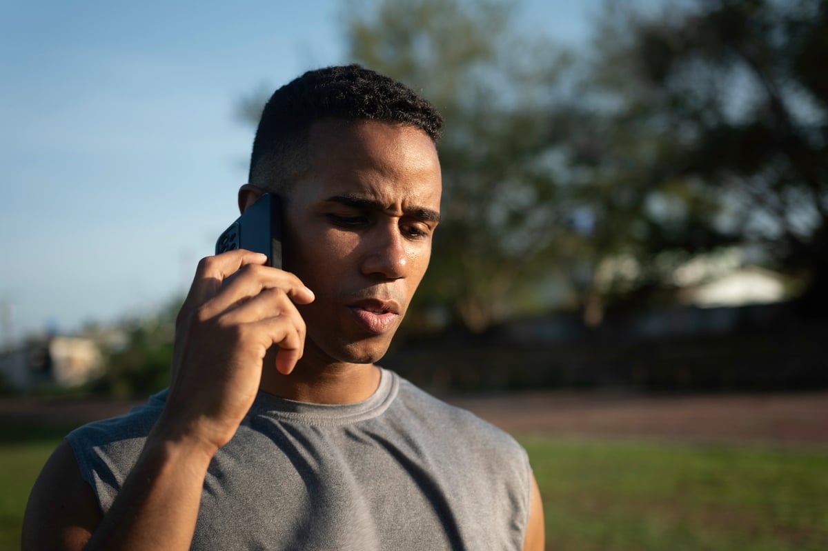 Young male athlete resting after an intense training session, engaging in a phone call while enjoying the warmth of the sun in a vibrant outdoor park setting