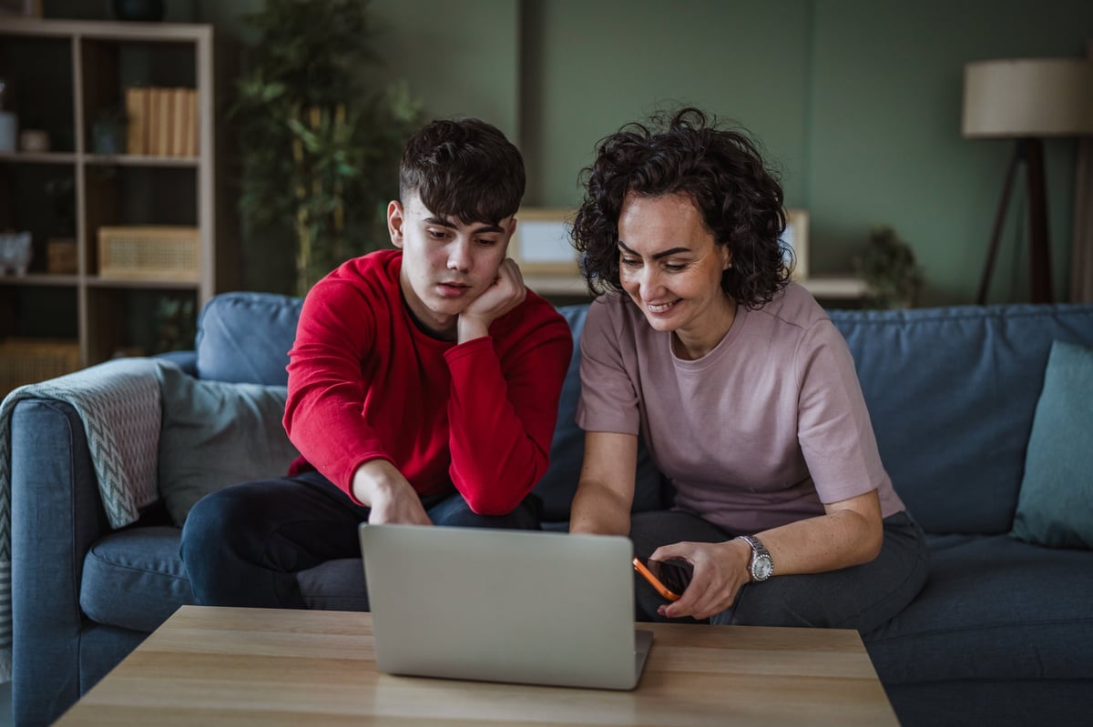 Mother and teenage son sitting on a sofa in the living room, sharing a laptop and discussing content, representing family connection, education, and digital lifestyle